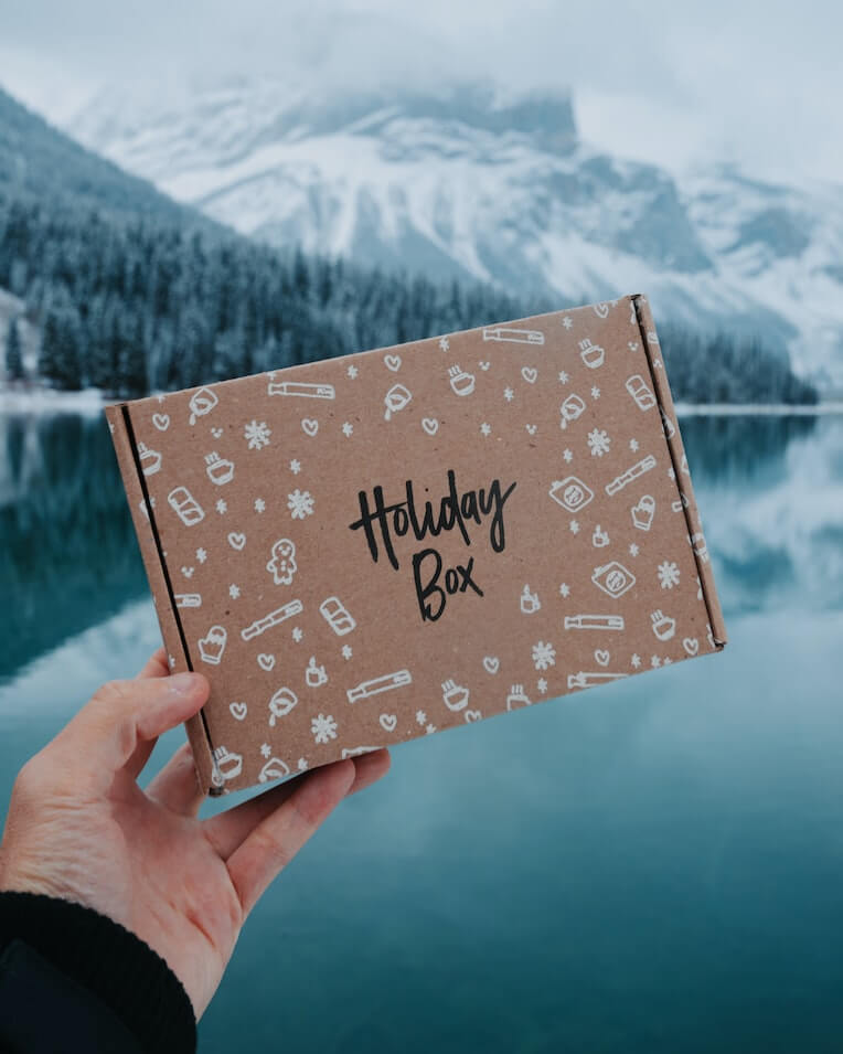 Hand holding a box labeled “Holiday Box” in front of a lake